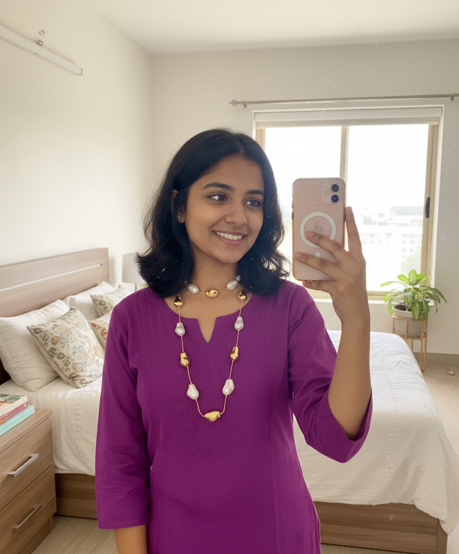 Woman taking a mirror selfie in a bedroom wearing a purple dress and white and gold necklace.
